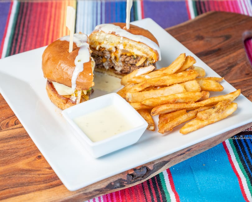 Two saucy sliders with cheese, french fries, and dipping sauce on a white plate over a colorful blanket.