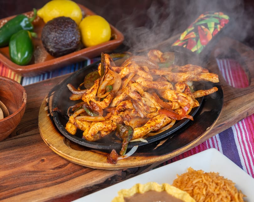 Sizzling chicken fajitas with peppers and onions, steam rising, served with lemons, avocado, and jalapeños.