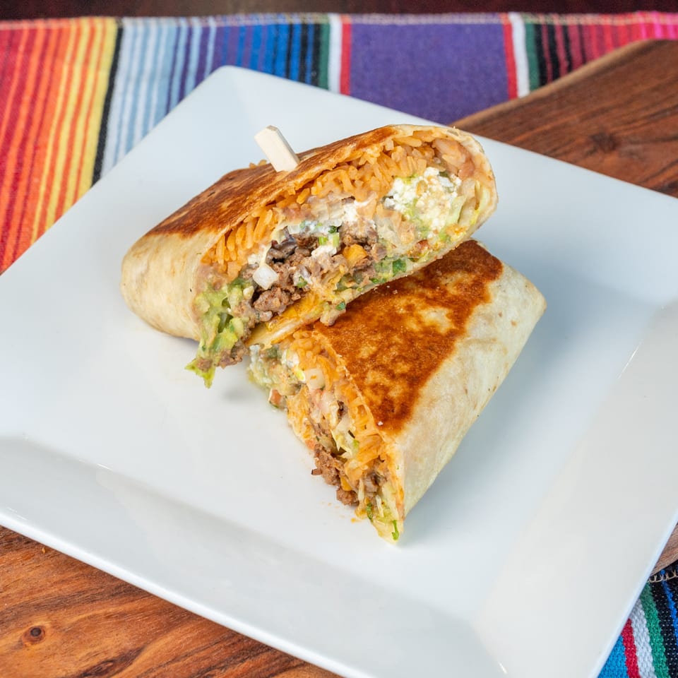 Grilled burrito cut in half, showing rice, ground meat, cheese, and guacamole on a white plate.