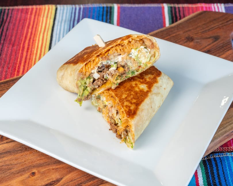 Grilled burrito cut in half, showing rice, ground meat, cheese, and guacamole on a white plate.