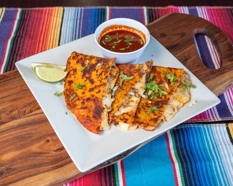 Cheesy birria tacos with cilantro, onion, lime, and dipping sauce, served on a colorful striped blanket.