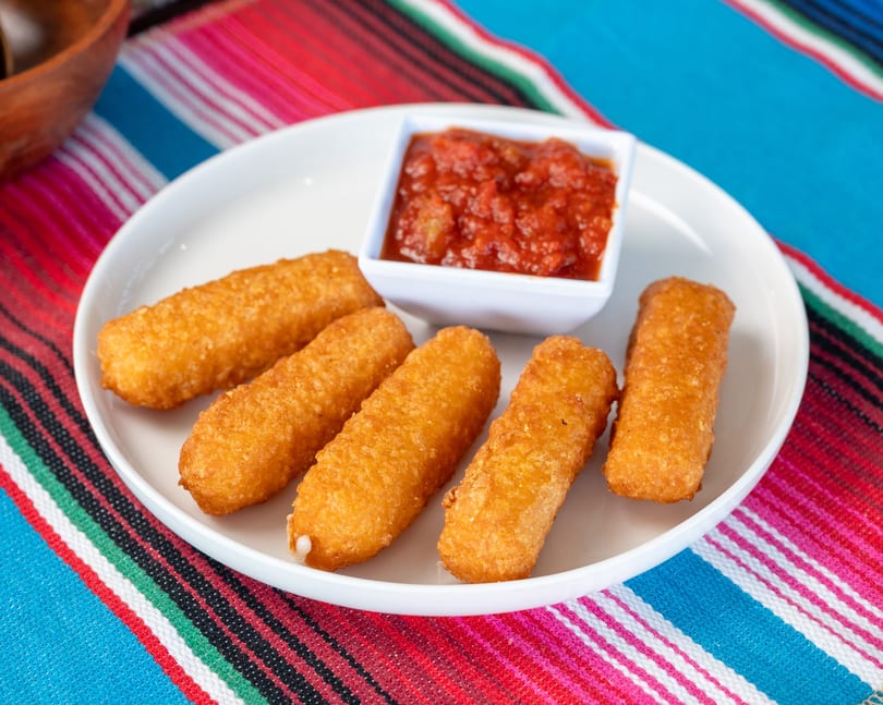 Crispy mozzarella sticks with marinara sauce on a white plate over a vibrant striped tablecloth.