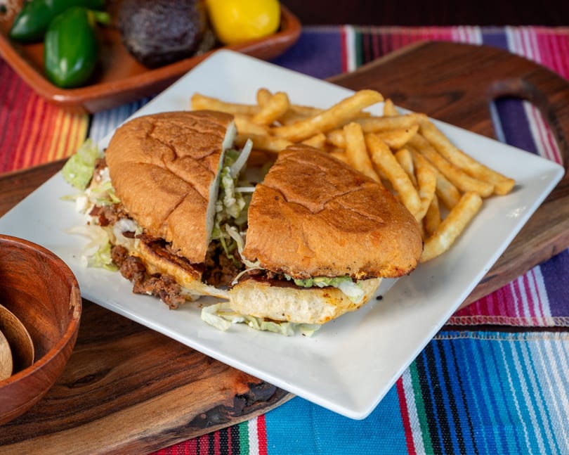 Mexican torta sandwich cut in half with golden fries on a white plate, on a colorful striped tablecloth.