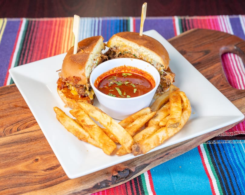 Birria sandwiches, golden fries, and red dipping sauce on a white plate over a colorful striped blanket.