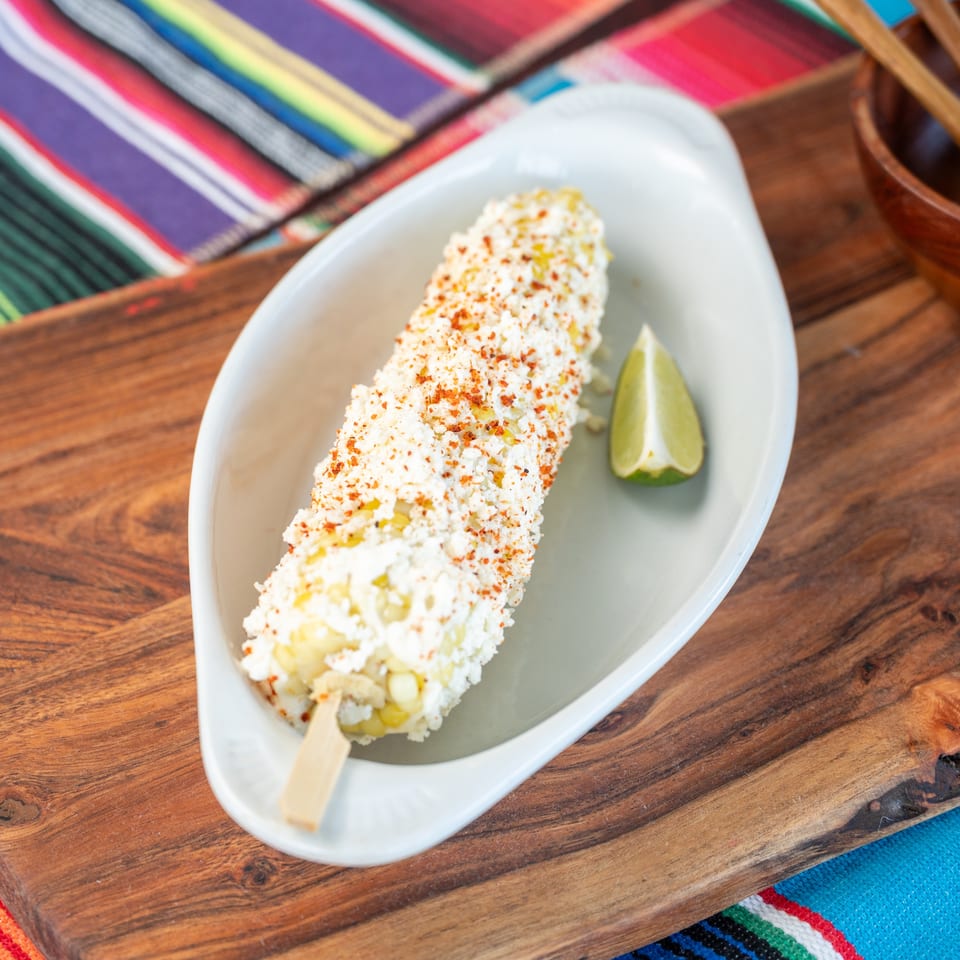 Elote on a stick, covered in white cheese and red spice, with a lime wedge.
