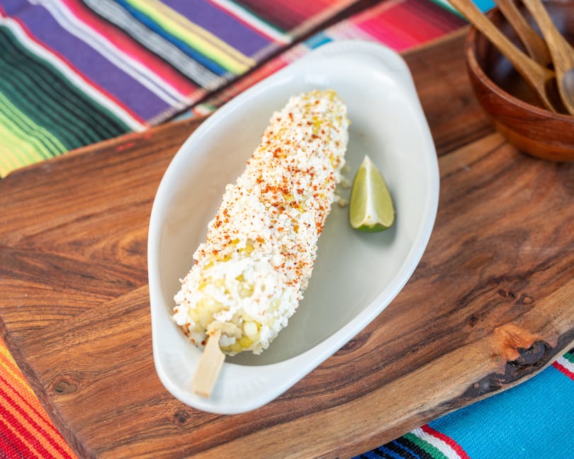 Elote on a stick, covered in white cheese and red spice, with a lime wedge.