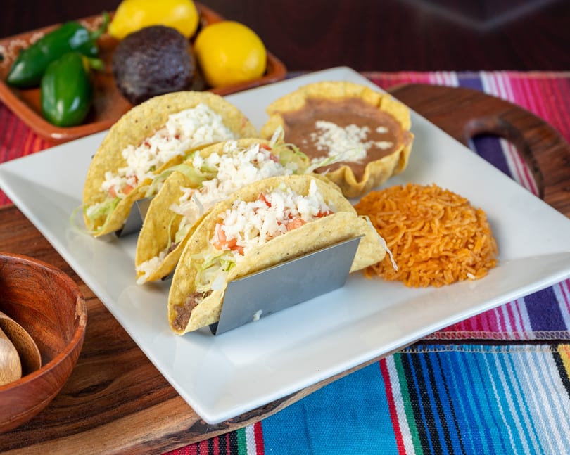 Three hard-shell tacos, Mexican rice, and refried beans on a white plate with a colorful striped tablecloth.